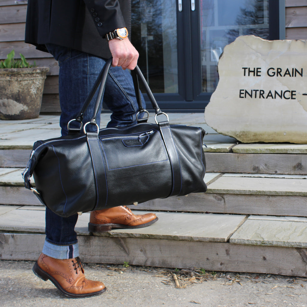 This  travel & duffel bag in black leather with blue stitching  Handcrafted in England from full grain calf leather, handmade smooth top handles, a removable and adjustable shoulder strap, solid nickel hardware Fully Harris Tweed lining Free UK shipping 